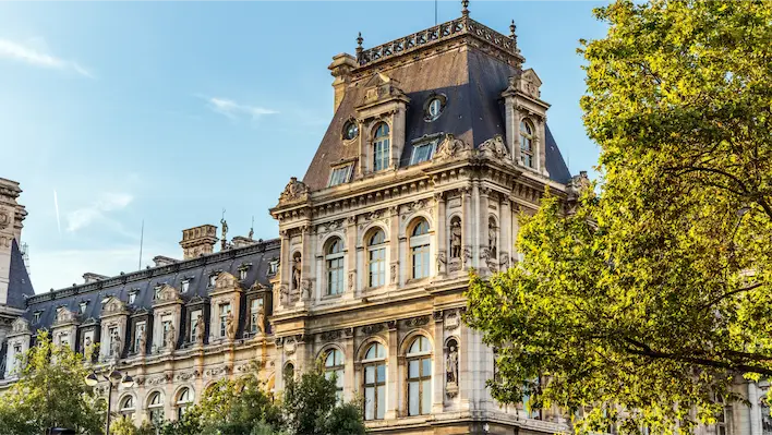 Façade d’un bâtiment historique en pierre avec toit mansardé et arbres au premier plan.